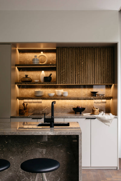 A modern kitchen featuring dark wood cabinets finished with Oil Plus 2C. An island with a stone water fall edge is in the  foreground and simple barstools are stationed at it.