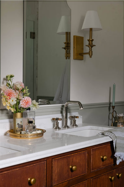 A renovated bathroom with a white oak vanity, brass wall sconces, marble countertop and more modern fixtures.