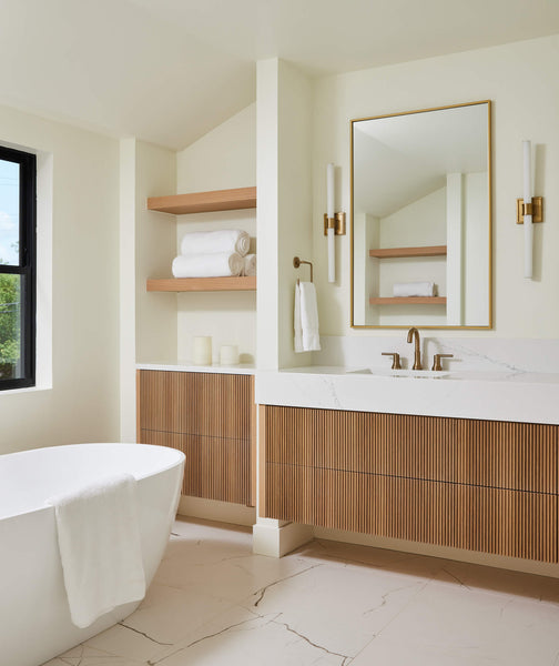 Light and airy bathroom with a floating slatted white oak vanity, white countertops and flooring, and brass fixtures. 